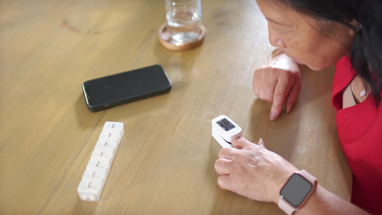 Senior Asian woman in red top after cell alert tapping white device, checking pillbox, copy space