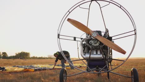 the motor paraglider stands in the field at sunset with a wooden propeller, and the pilot lays out the parachute and aligns the slings