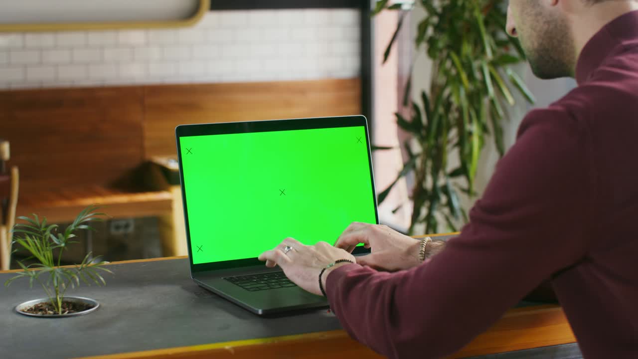 un joven está escribiendo en una computadora portátil con una pantalla verde sentado en una cafetería