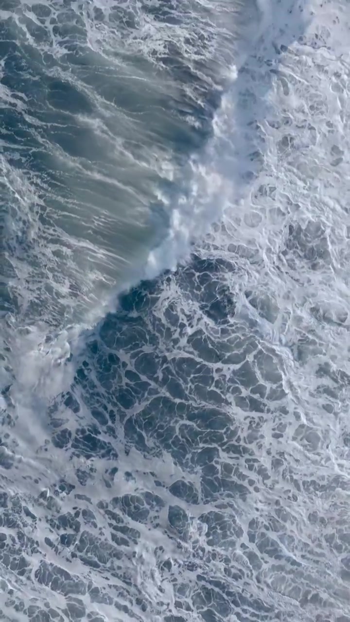 Straight-down drone view of ocean waves and white sea foam washing over the black volcanic sands of Khalaktyrsky Beach on the Kamchatka Peninsula, Russia, creating a dramatic coastal texture