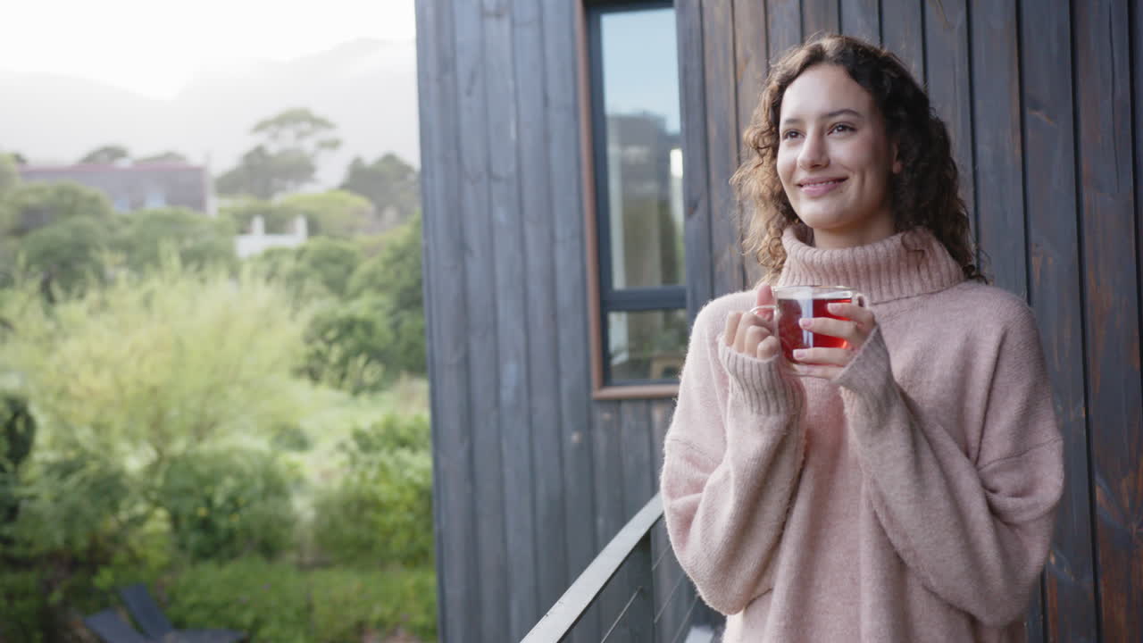 Smiling woman holding cup of tea, relaxing on balcony in cozy sweater, copy space