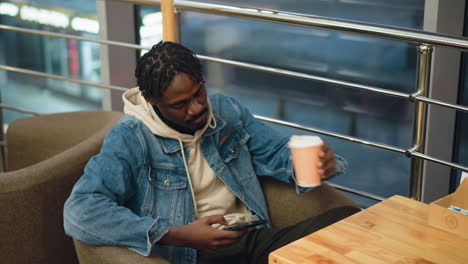 young man in denim jacket and beige hoodie leans back in soft chair, holding smartphone in one hand and takeaway coffee cup in other, relaxing in cozy cafe with city lights visible through large window