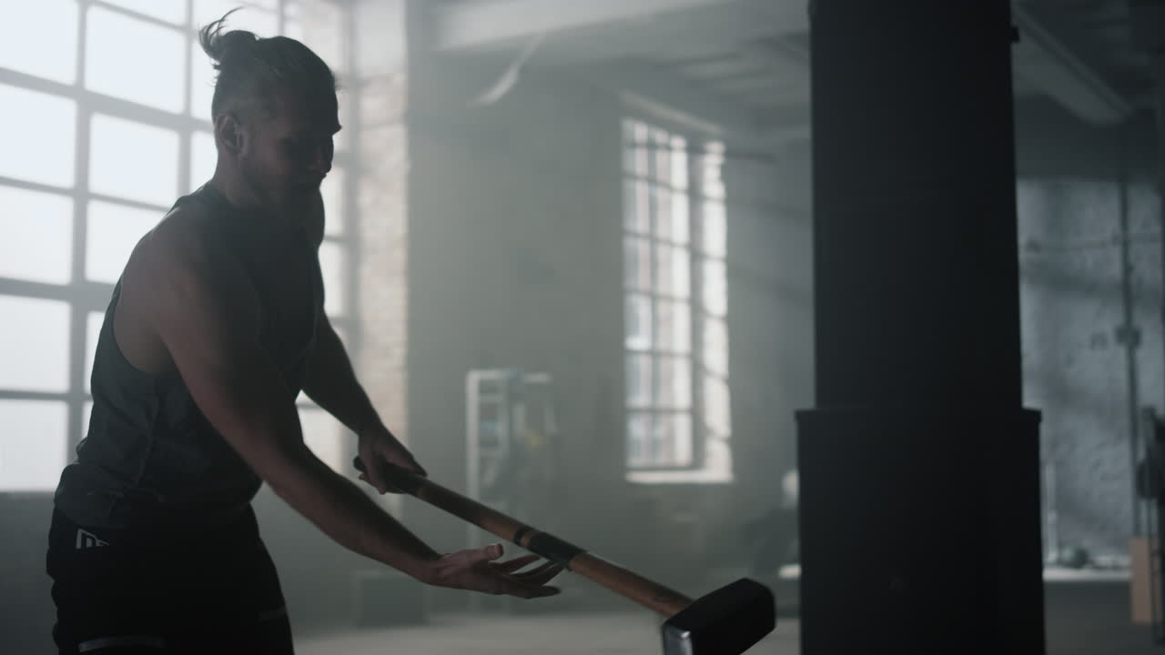 화난 남자가 망치로 크로스피트 타이어를 때리고 있다. 남자가 신체 운동을 하고 있다.