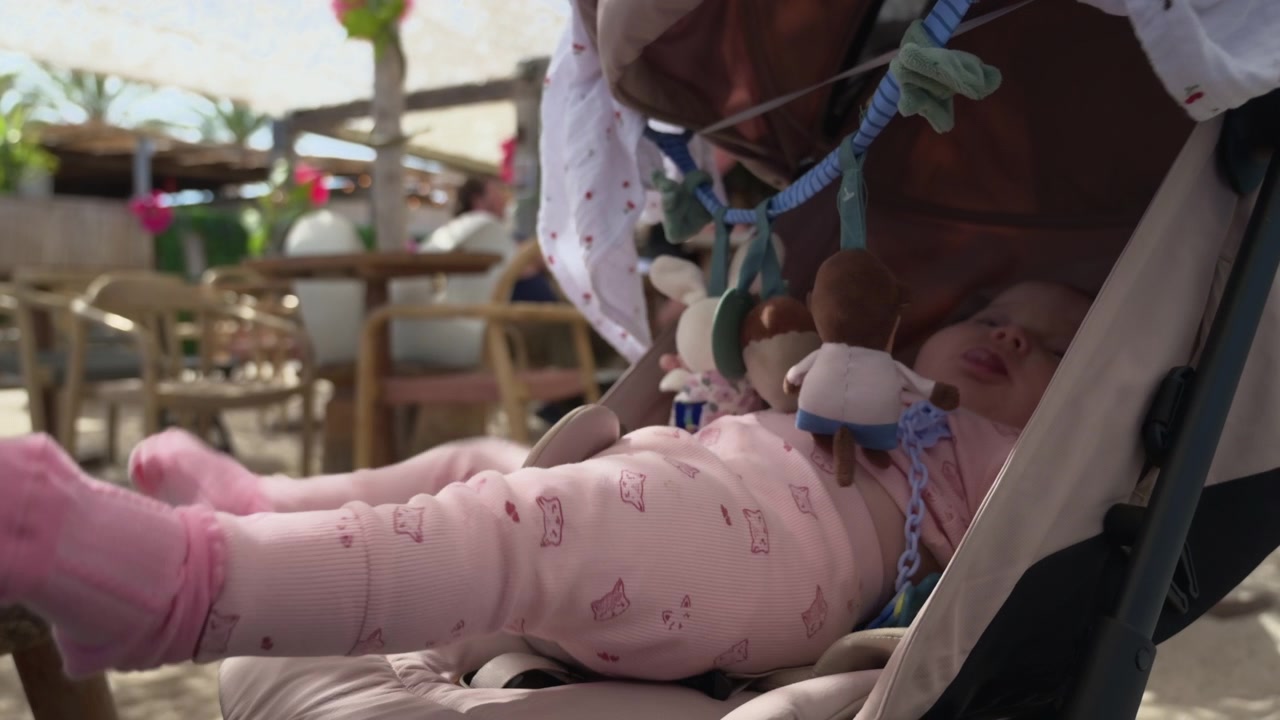 Baby in a stroller at a beach club with toys hanging overhead