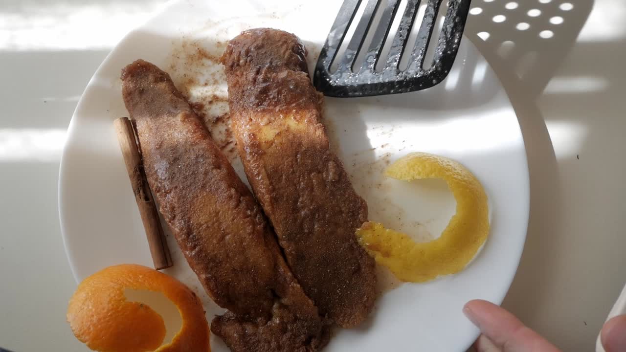 Torrijas on a plate, cinnamon sprinkled on top, with orange peels and a cinnamon stick garnish