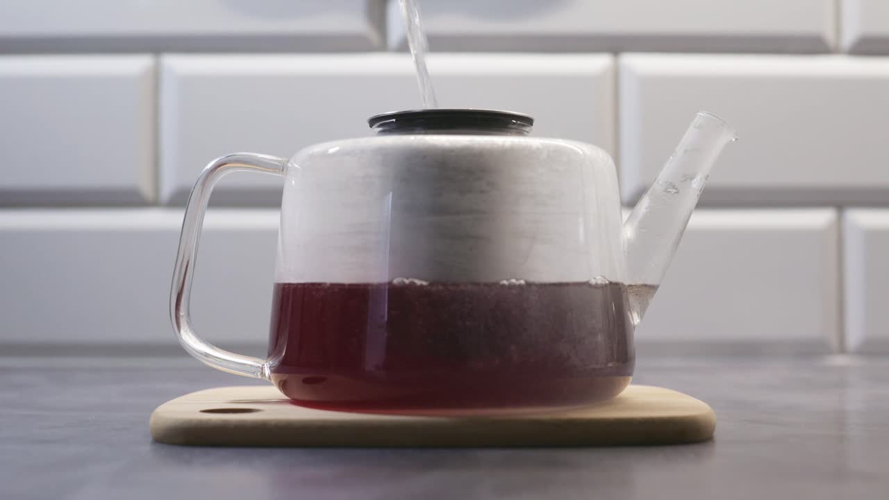 Teiera minimalista in vetro con acqua calda versata all'interno, che prepara delicatamente il tè su un piano di lavoro della cucina. Il video trasmette calore, relax e un rito quotidiano del tè in un ambiente domestico moderno.