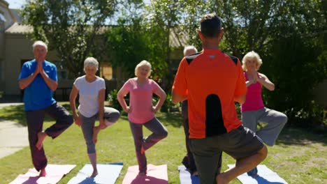 Vista-Trasera-De-Un-Entrenador-Masculino-Caucásico-Entrenando-A-Personas-Mayores-En-La-Realización-De-Yoga-En-El-Jardín-De-N-4k