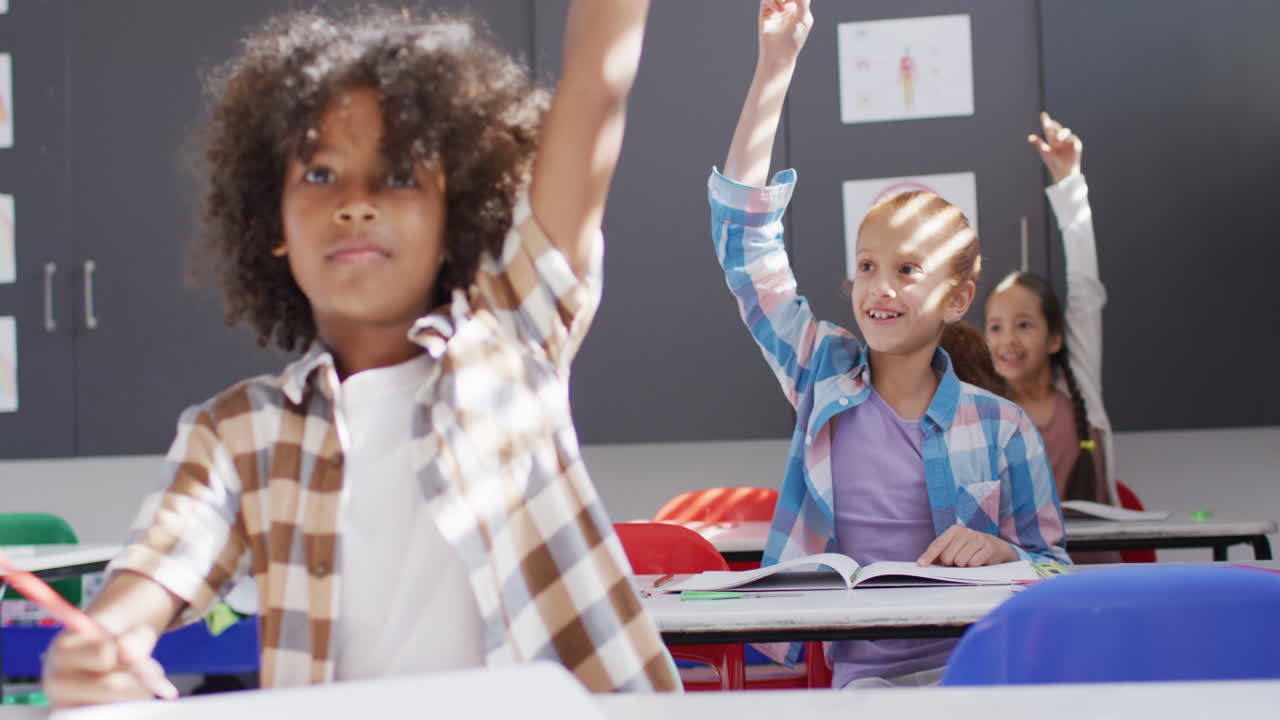 Diverse happy schoolchildren sitting at desk and raising hands in school classroom