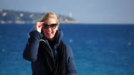 a young blonde woman laughs to the camera and puts her sunglasses on, the blue sea in soft focus behind her