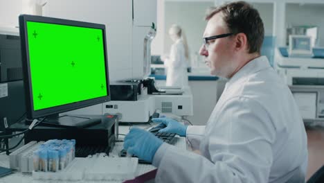 male research scientist sits at his workplace in laboratory, uses green mock-up screen personal computer. i the background genetics, pharmaceutical research centre with innovative equipment.