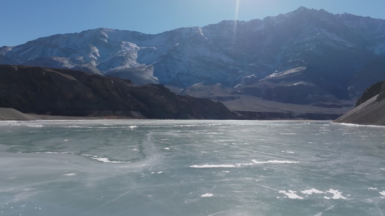Die gefrorene Oberfläche des Indus mit Eismustern erstreckt sich im Vordergrund unter kahlen Himalaya-Gipfeln nahe dem Dorf Basgo. Eine Weitwinkel-Luftaufnahme fängt die hochalpine Winterlandschaft ein.