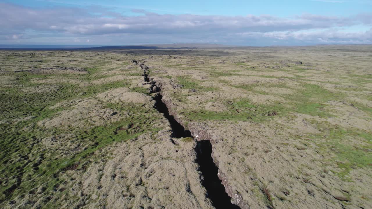 Aerial view of a large crack in the ground between two tectonic plates in a volcanic rift zone in Iceland. The landscape shows the distinct separation of the plates and the rugged terrain.