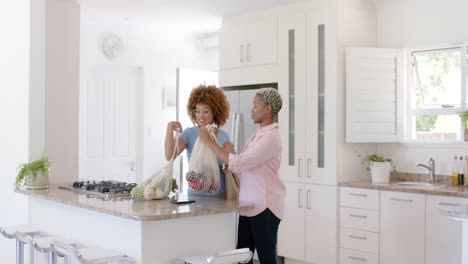 happy diverse female lesbian couple holding bags with groceries in kitchen in slow motion