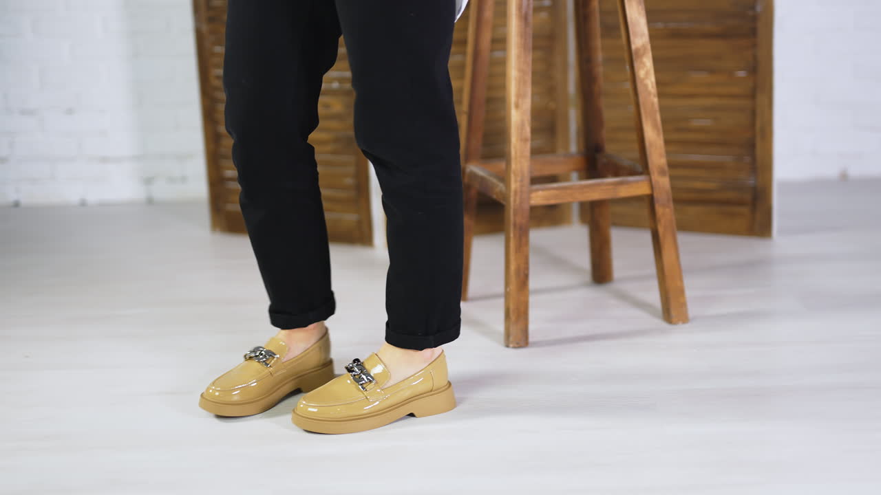 Female walking back and forth in fashionable new shoes. Beige stylish footwear on plain soles with chain decoration. Wooden stool and folding screen at backdrop.