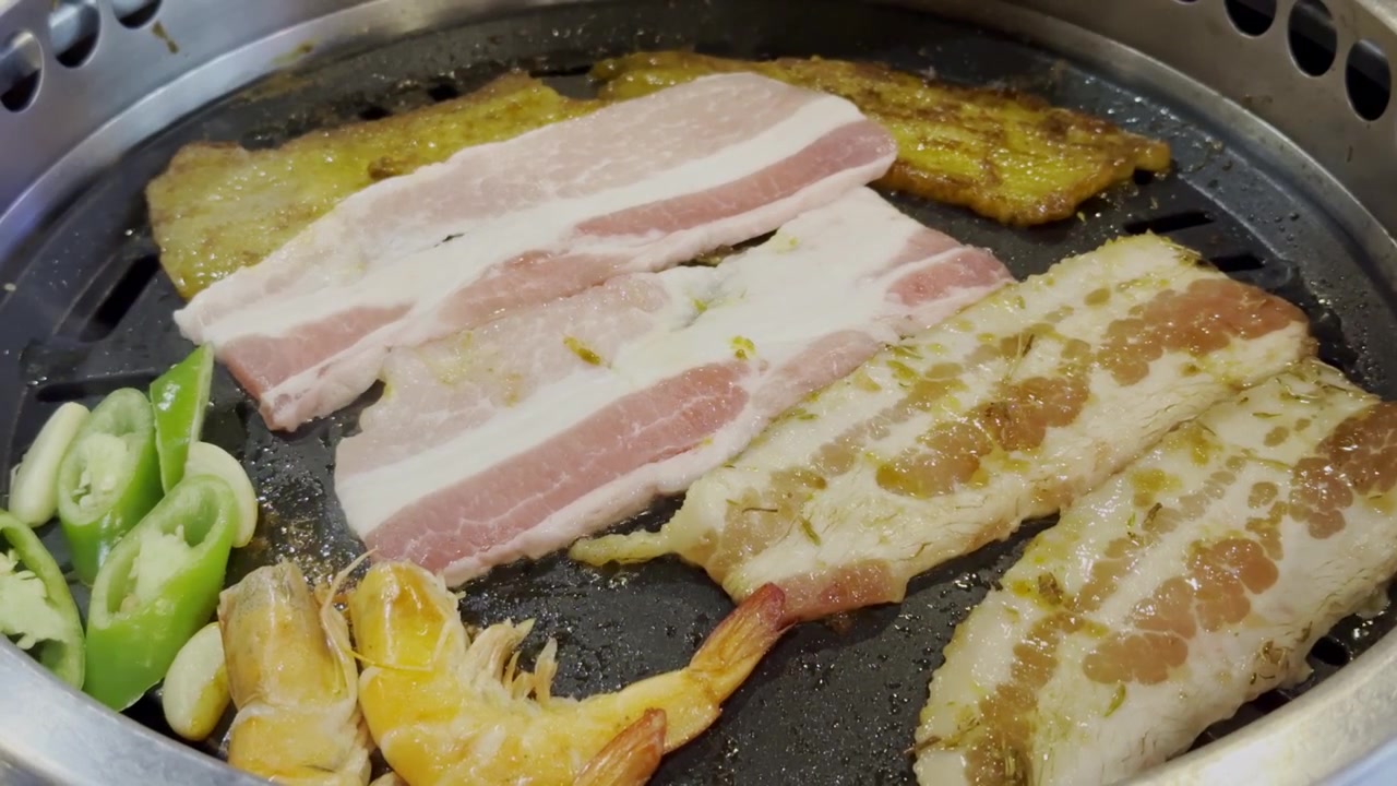 伝統的な食事の場面で、熱い韓国式焼肉プレートで焼かれる薄切り豚肉のクローズアップ。ジュージューという音、立ち上る湯気、そして美味しい風味が特徴。