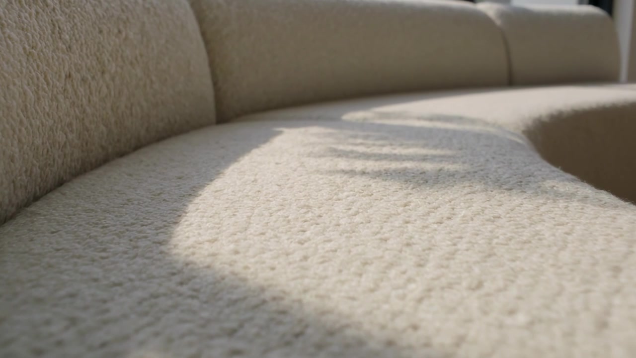 Luce solare che si sposta dalla finestra attraversando il sedile di un divano beige, proiettando un'ombra di foglia su tessuto testurizzato a casa.