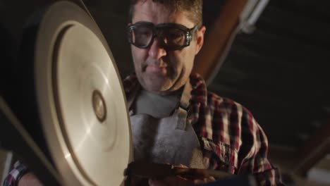 close up of caucasian male knife maker in workshop wearing glasses and using sander