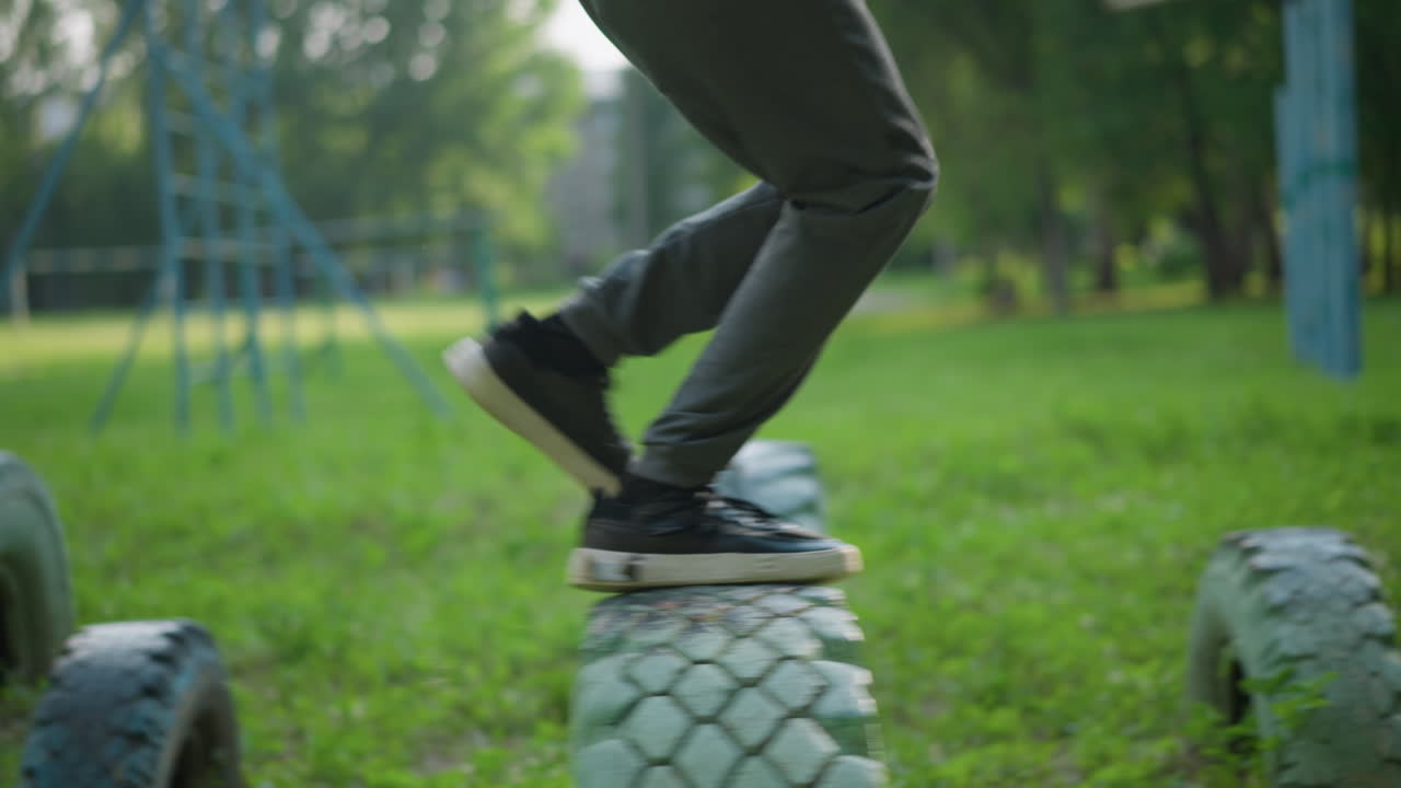 Close-up of a person in gray trousers and sneakers sprinting across three tires in a grassy field, jumping over two with blurred goalposts and trees in the background