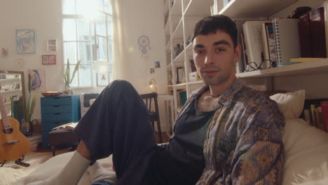 young man sitting on bed and posing for camera in sunlit room