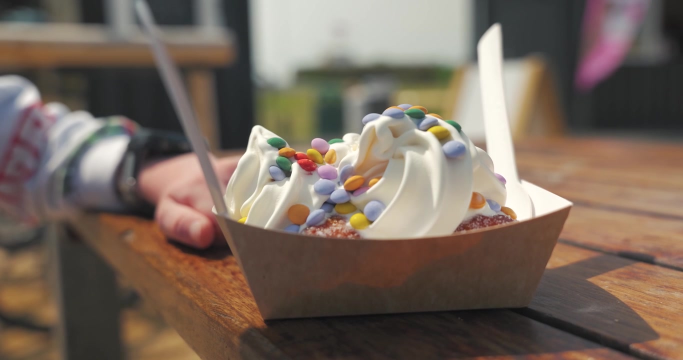 Slow motion close up of a donut dessert topped with ice cream and colourful sprinkles on an outdoor table near Littlehampton seafront