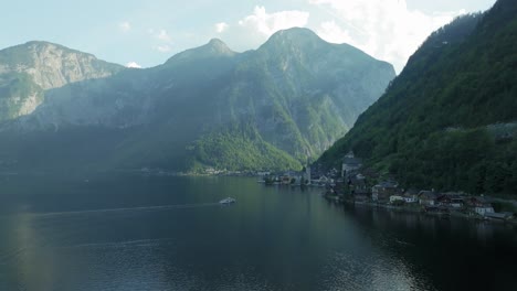 Drone-glides-forward-over-the-stunning-lake-towards-Hallstatt-village,-nestled-amidst-colossal-mountains-that-throw-it-into-the-afternoon-shadow,-as-cruise-boats-approach-the-docks