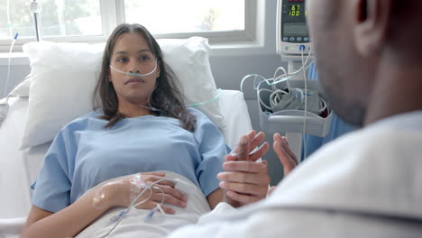 un médico masculino diverso sosteniendo la mano de una paciente con un oxímetro en la cama del hospital, en cámara lenta