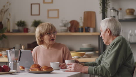 elderly couple using laptop and speaking over tea at kitchen table