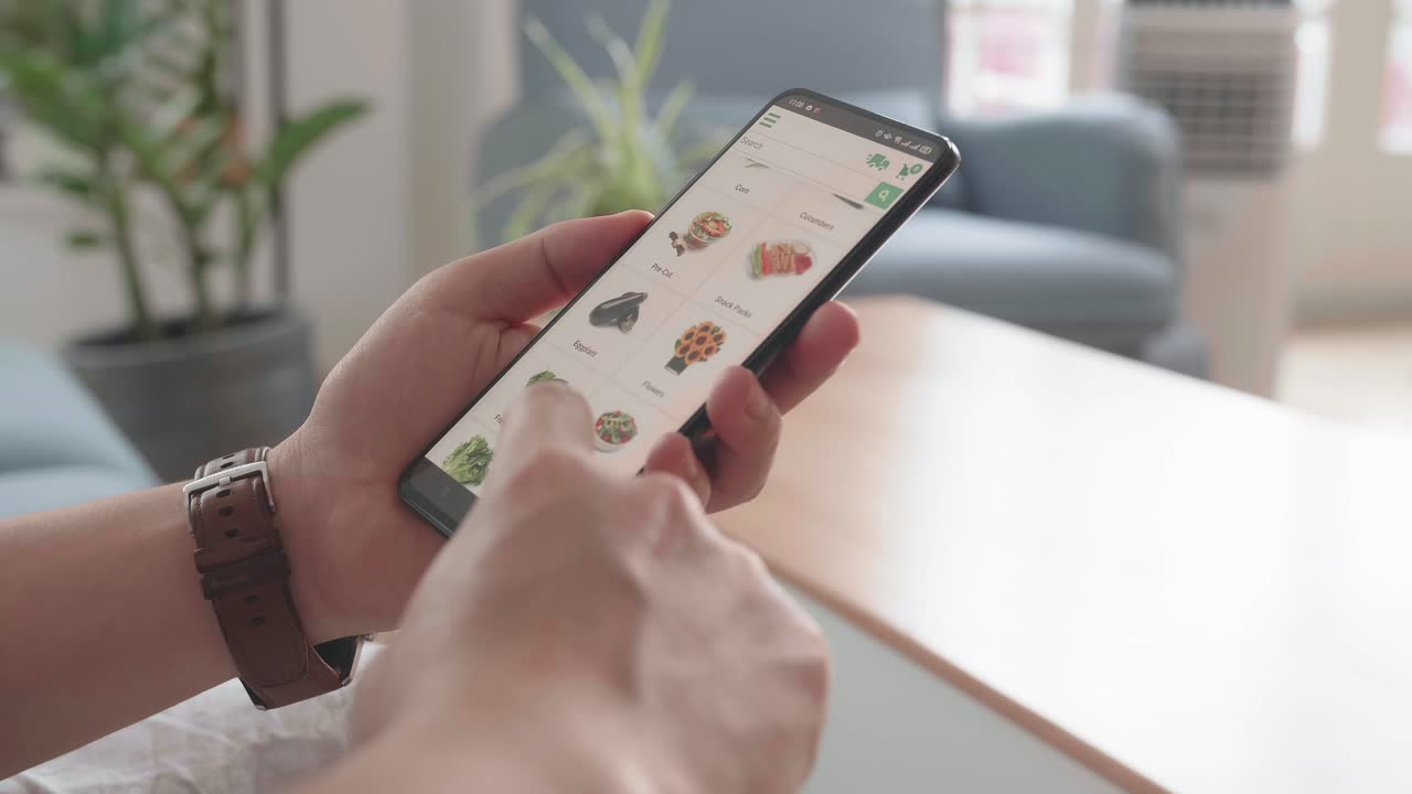 Man Sitting On Couch Using Smartphone Buys In Internet Shop. Male Selects Vegetables In Grocery Online Store