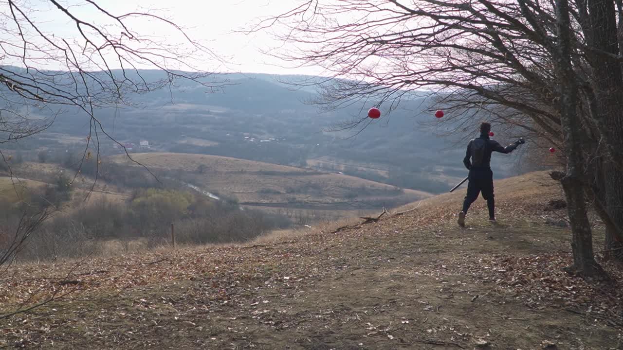 A stealth ninja clad in dark attire strides along a rugged dirt path as red spherical targets dangle from tree branches against rolling hills. perfect visual for martial arts.