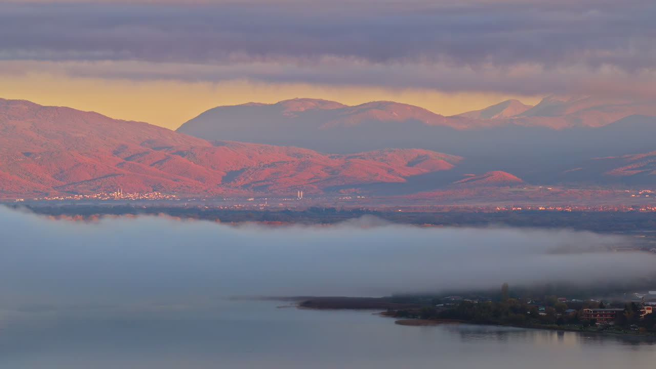 Aerial drone view of the Ohrid, Macedonia region, showing fog lifting from the wetlands and soft pink light falling on the surrounding mountains and villages