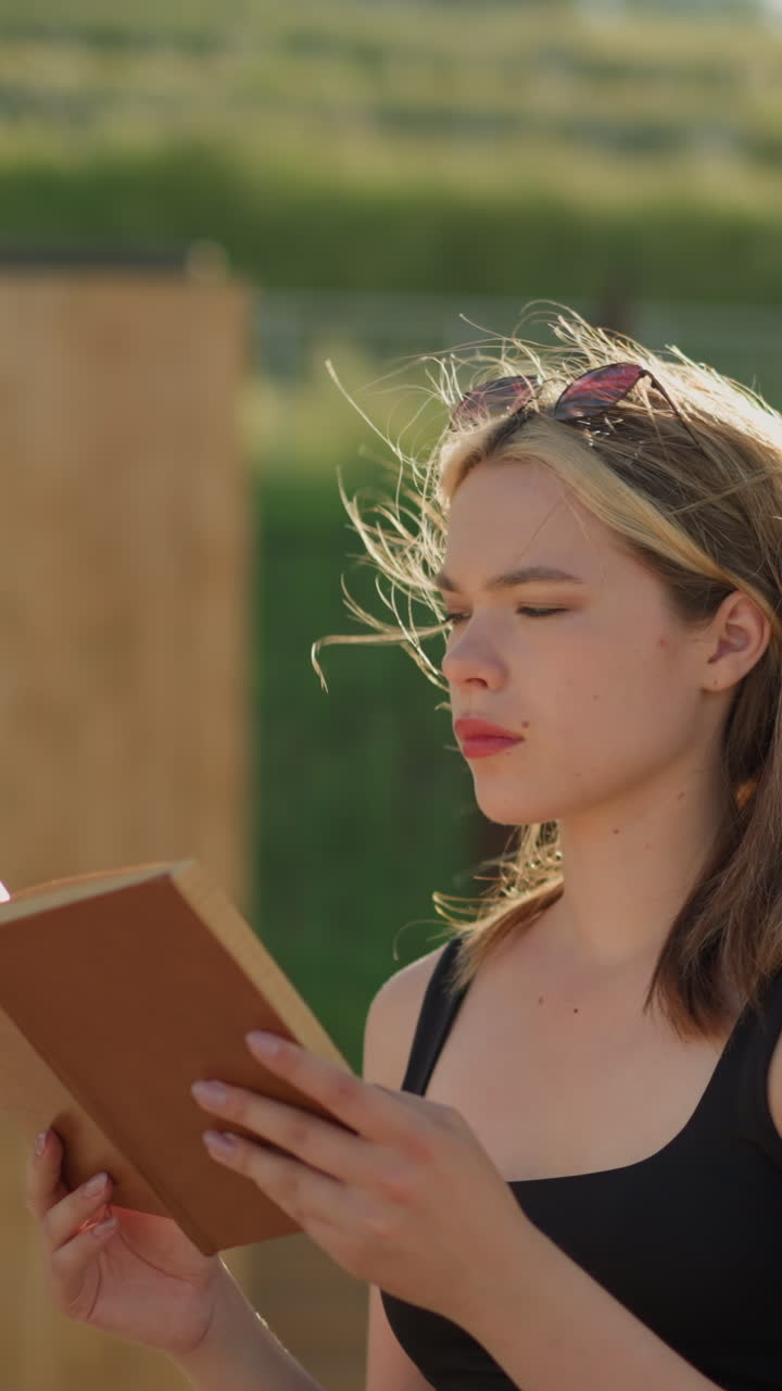 mujer sentada, leyendo un libro mientras el viento sopla suavemente su cabello, la luz del sol la ilumina mientras lee, con una colina de vegetación exuberante y una estructura de madera borrosa en el fondo