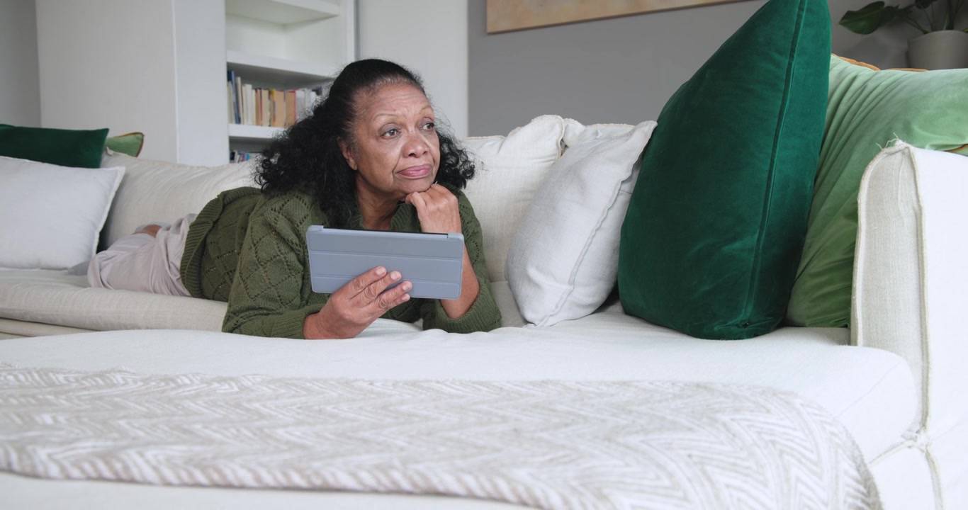 Senior African American woman in olive green sweater using gray tablet tapping, reading at home