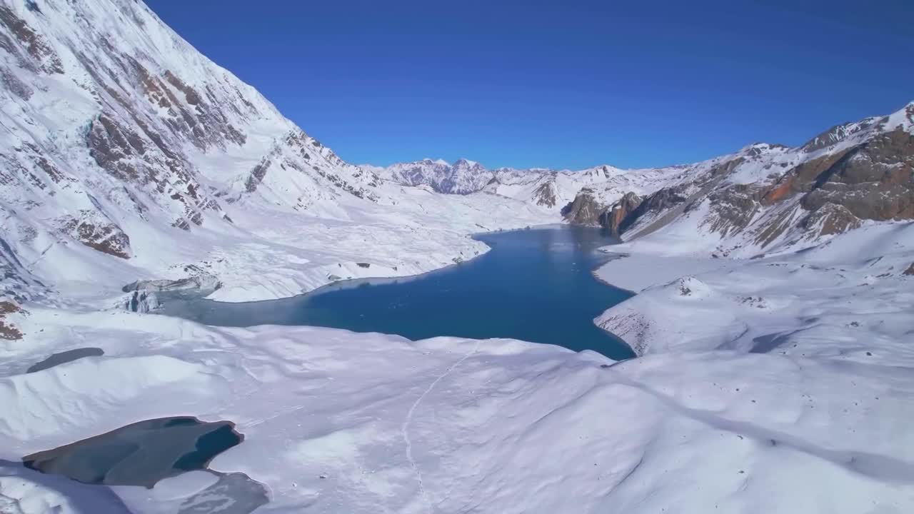 Ascending aerial drone view panorama of Tilicho Lake in Nepal, Annapurna Circuit trekking route. Clear blue glacial lake is surrounded by snowy mountains ranges during sunny day with clear blue sky