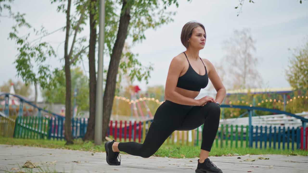 Kvinde strækker ben i en forlystelsespark, udfører lunges, mens en rutsjebane bevæger sig i baggrunden, livlig legeplads og udendørs aktiviteter, viser en aktiv livsstil og fitnessrutine.