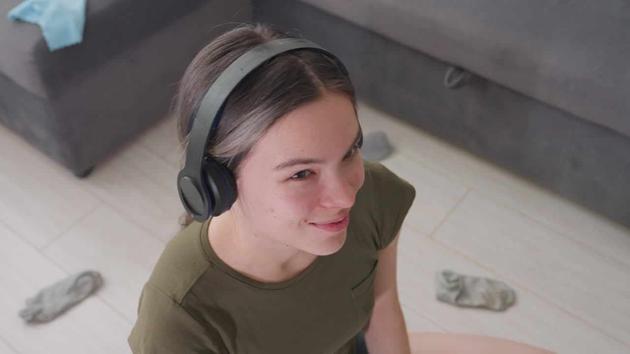 Young woman with headphones nodding head to rhythm of music while looking upward with relaxed smile, seated indoors in casual wear, natural daylight