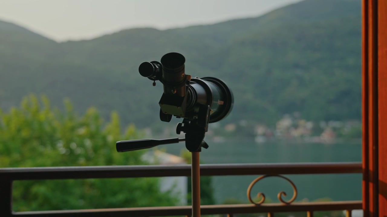 Nærbillede af et teleskop på et stativ ved siden af en balkon med udsigt over en fredelig bjergsø, oplyst af blødt morgenlys. Indbydende alpeferie og naturskønt udsigtspunkt.