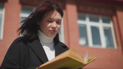 primer plano de un estudiante leyendo al aire libre mientras da la vuelta a las páginas de un libro, el cabello revoloteando en el viento suave con un edificio de ladrillo rojo en el fondo bajo la luz del sol brillante