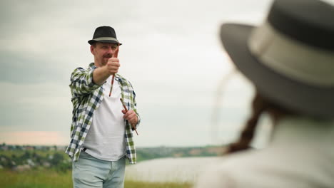 a cheerful painter wearing a checked shirt and holding a brush smiles as a blurred woman is visible in the background. the scene captures a joyful and creative moment in an outdoor setting