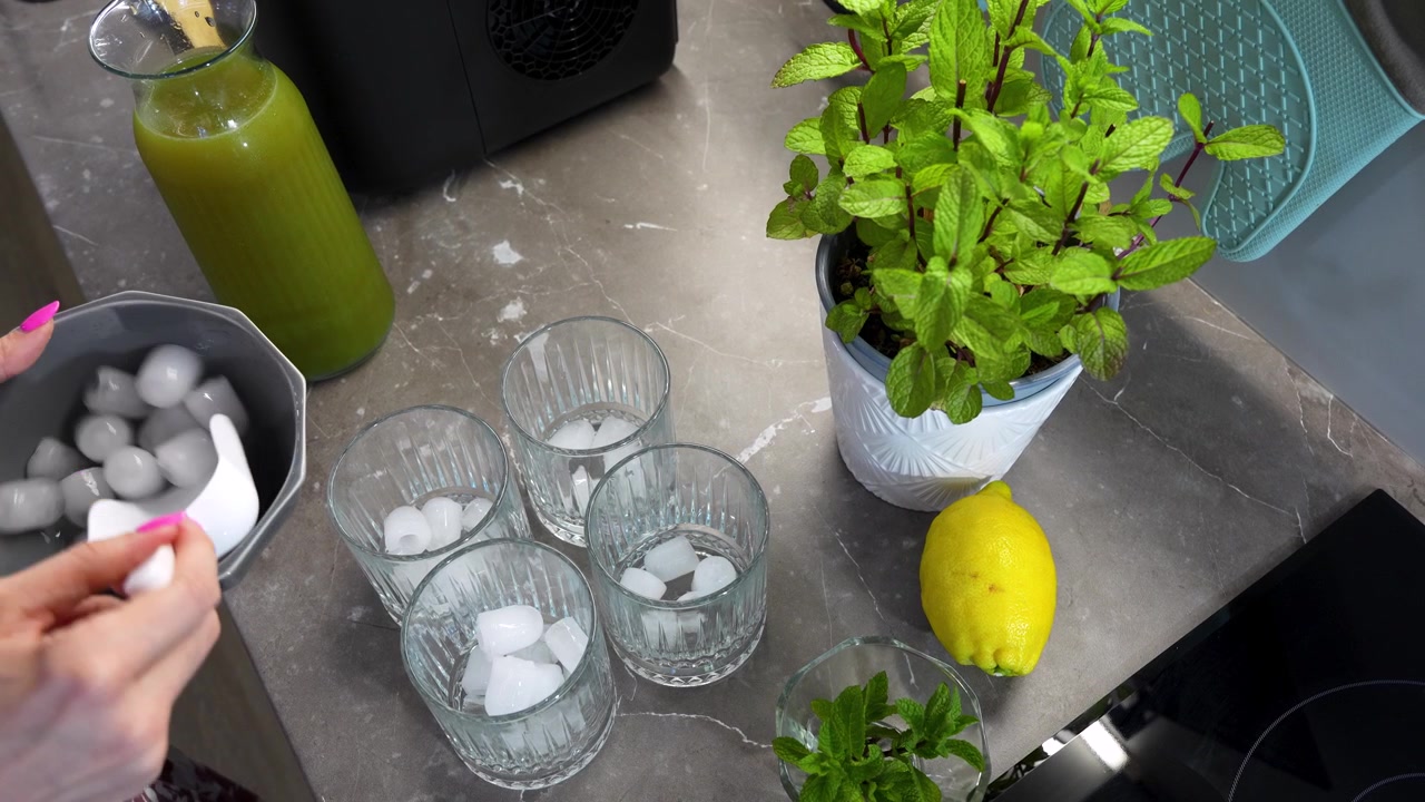 Kädet asettavat jääpaloja laseihin marmorisella työtasolla tuoreiden minttukasvien ja sitruunan vieressä, valmistellen virkistävää arabialaista kesäistä minttulimonadia.