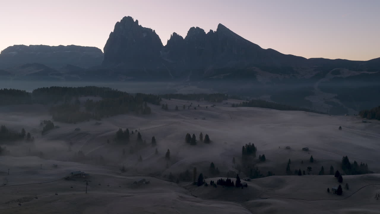 Filmisk luftfoto af frostklædte Alpe di Siusi-enge med spredte bjerghytter og morgentåge, dramatiske Dolomitter-toppe i de italienske alper ved solopgang, fredfyldt natursceneri