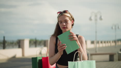 lady seated on outdoor bench with shopping bags on both sides retrieves a mint color book from mint green bag, opens it, and flips through its pages, while background remains slightly blurred