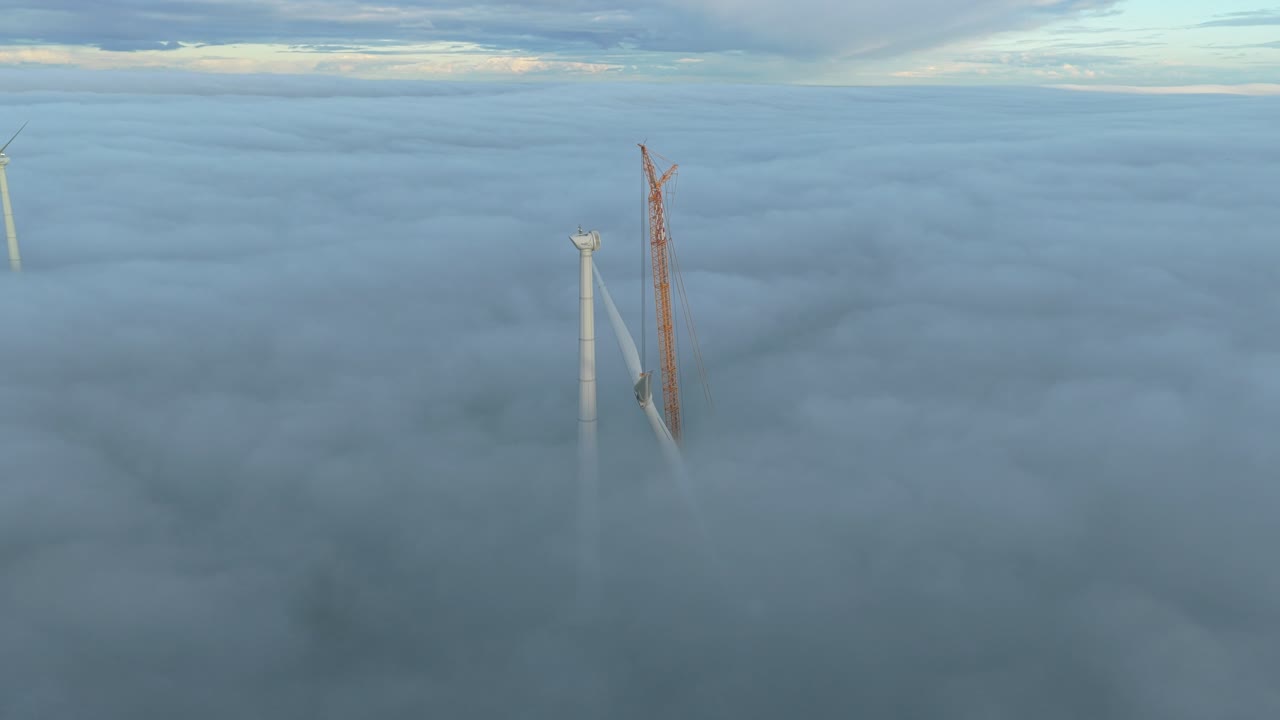 Wind turbine under construction as an orange crane mounts blades in the mist