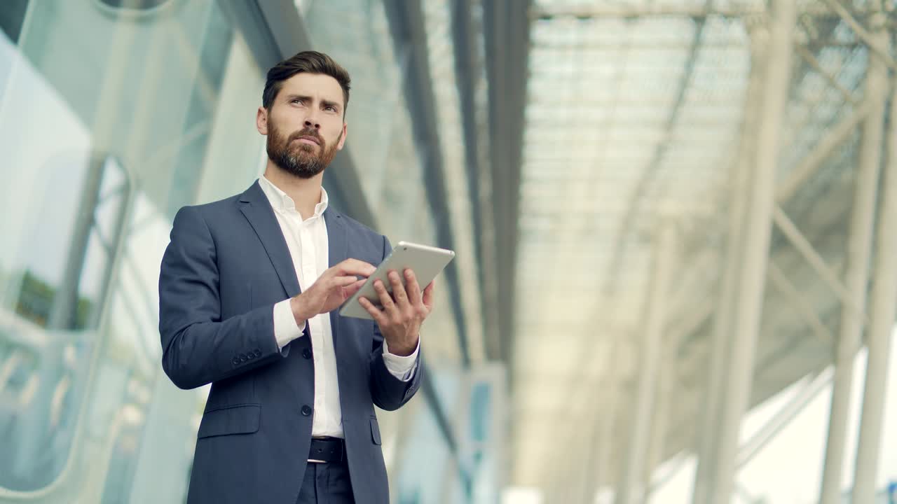 empresário barbudo elegante em terno de negócios formal em pé trabalhando com tablet nas mãos no fundo de um edifício de escritórios modernos do lado de fora.