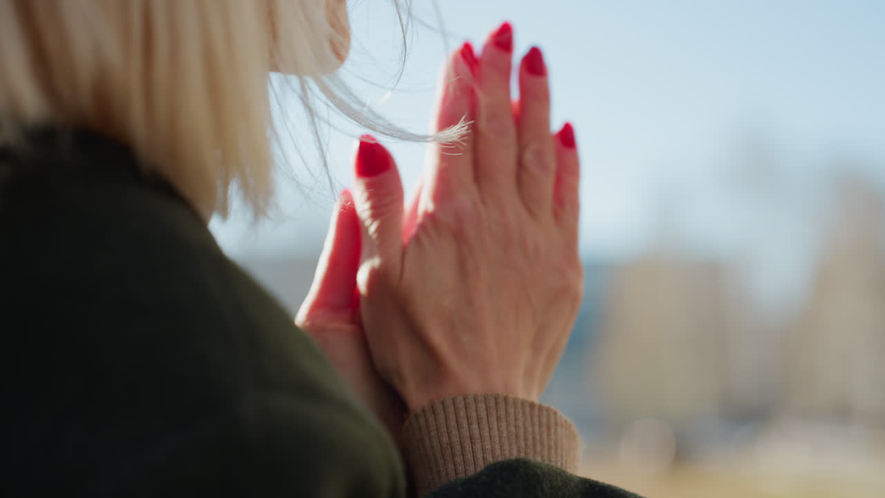 Back view of mature woman with blonde hair outdoors, wearing dark coat, holding hands with red nails together, sunlight highlighting strands, gesture suggesting gratitude in natural environment