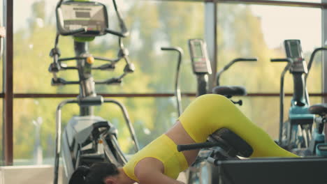 young hispanic woman in a yellow tracksuit performs a hyperextension exercise to train the muscles of the back. a brunette woman raises the body on bench in the gym to strengthen the back and posture