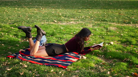 eine wunderschöne junge frau, die ein buch liest und auf einem grasbewachsenen parkfeld lernt, während herbstblätter im wind in zeitlupe wehen