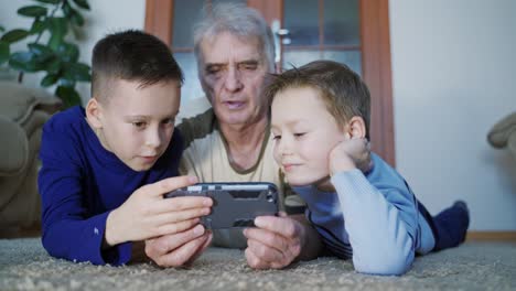 old man with boys playing video game. teenage boys typing on the smartphone while lying on the floor with grandpa at home