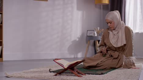 muslim woman wearing hijab praying with prayer beads at home on prayer mat with open copy of the quran on stand