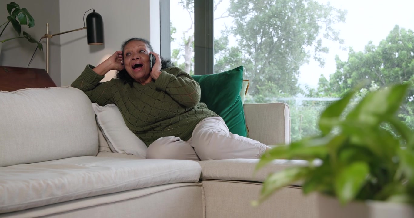 Mulher afro-americana idosa lendo smartphone, atendendo a uma chamada, rindo no sofá, de suéter