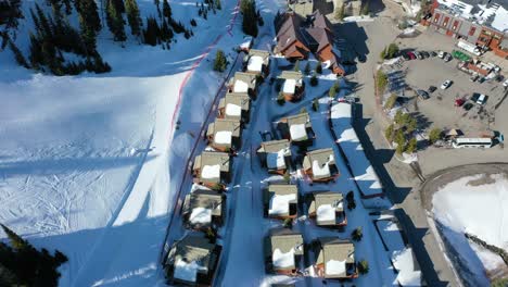 overhead drone footage of alpine vacation condos with snow-covered rooftops, nestled among trees and framed by mountains under a soft, glowing winter sky